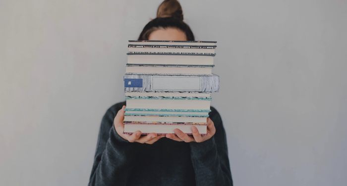 a fair skinned femme looking person holding a stack of books that is obscuring their face.jpg.optimal